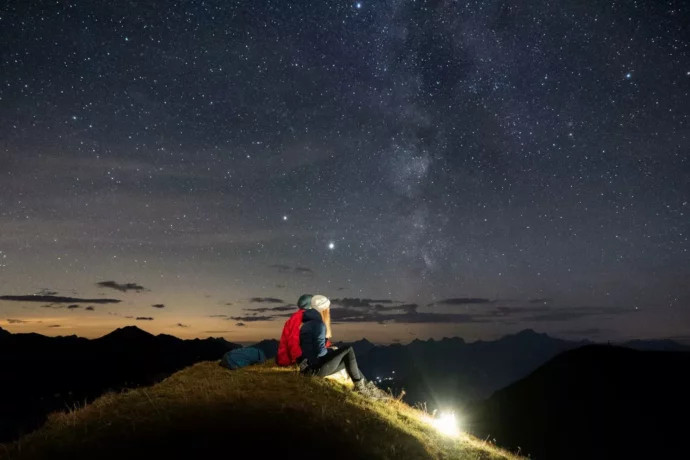 Zwei Personen sitzen auf einem Hügel und betrachten den Sternenhimmel.