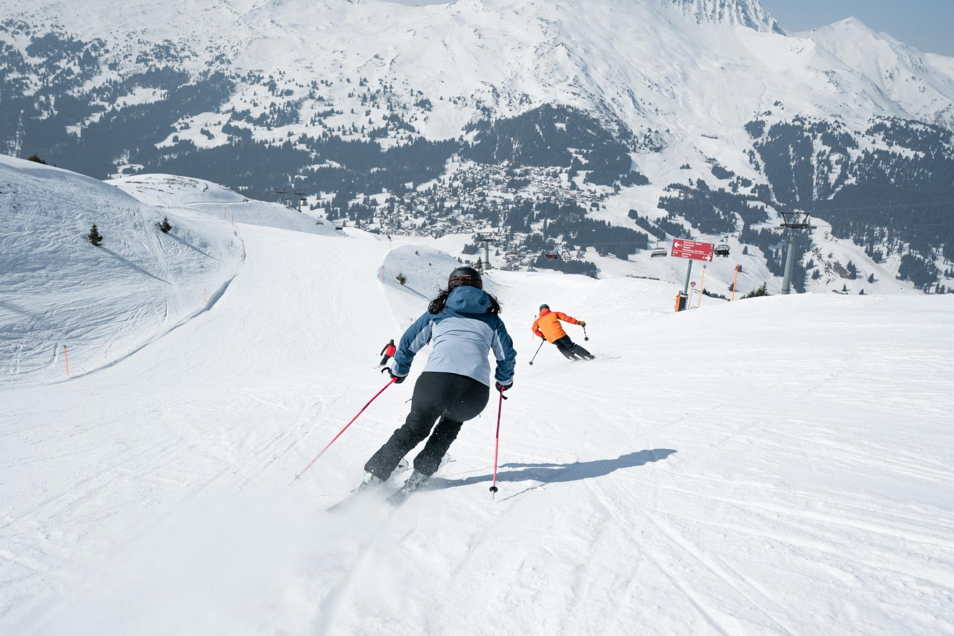 Drei Skifahrer fahren die Skipiste runter. Auf der gegenüberliegenden Seite sieht man die verschneiten Berge.