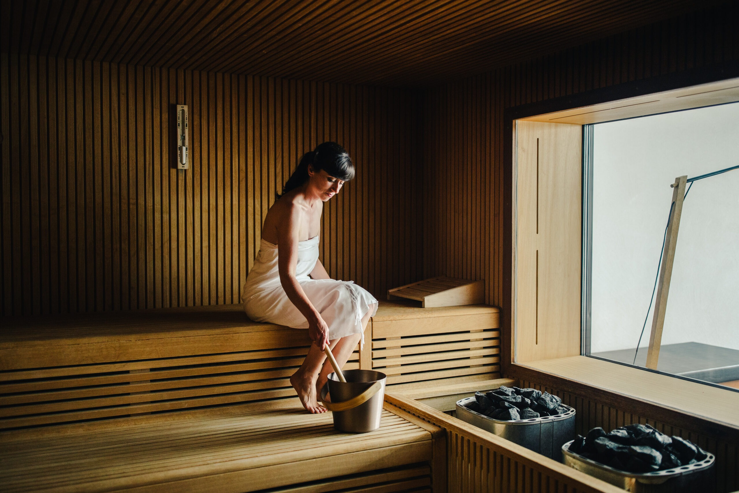 Frau sitzt in einer Sauna und giesst Wasser auf die heissen Steine.