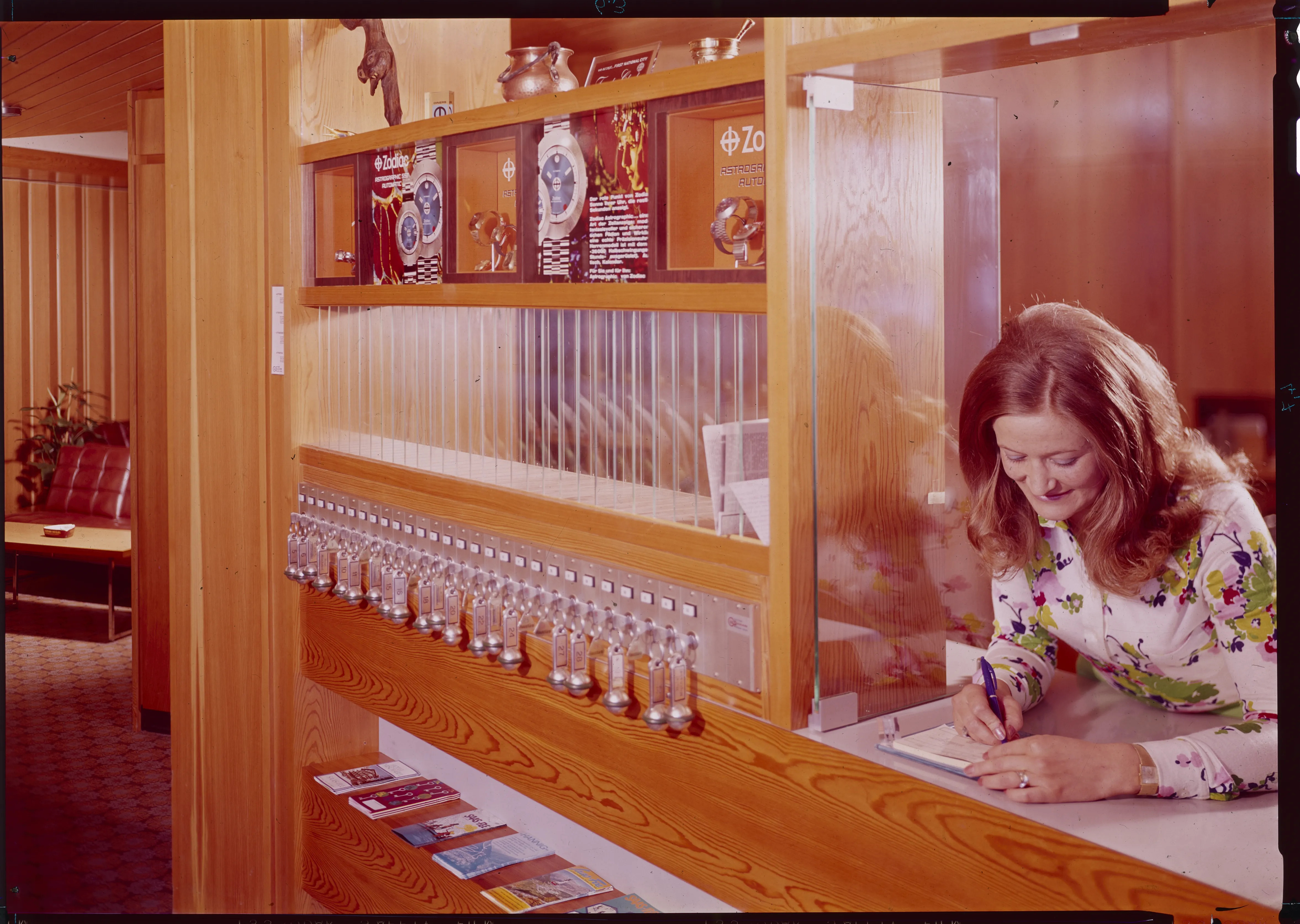 Rezeption im Retro-Stil mit Holzverkleidung, klassischen Hotelschlüsseln in Reih und Glied und einer Mitarbeiterin, die ein Formular ausfüllt; dahinter eine Glaswand und Auslagen mit Broschüren.