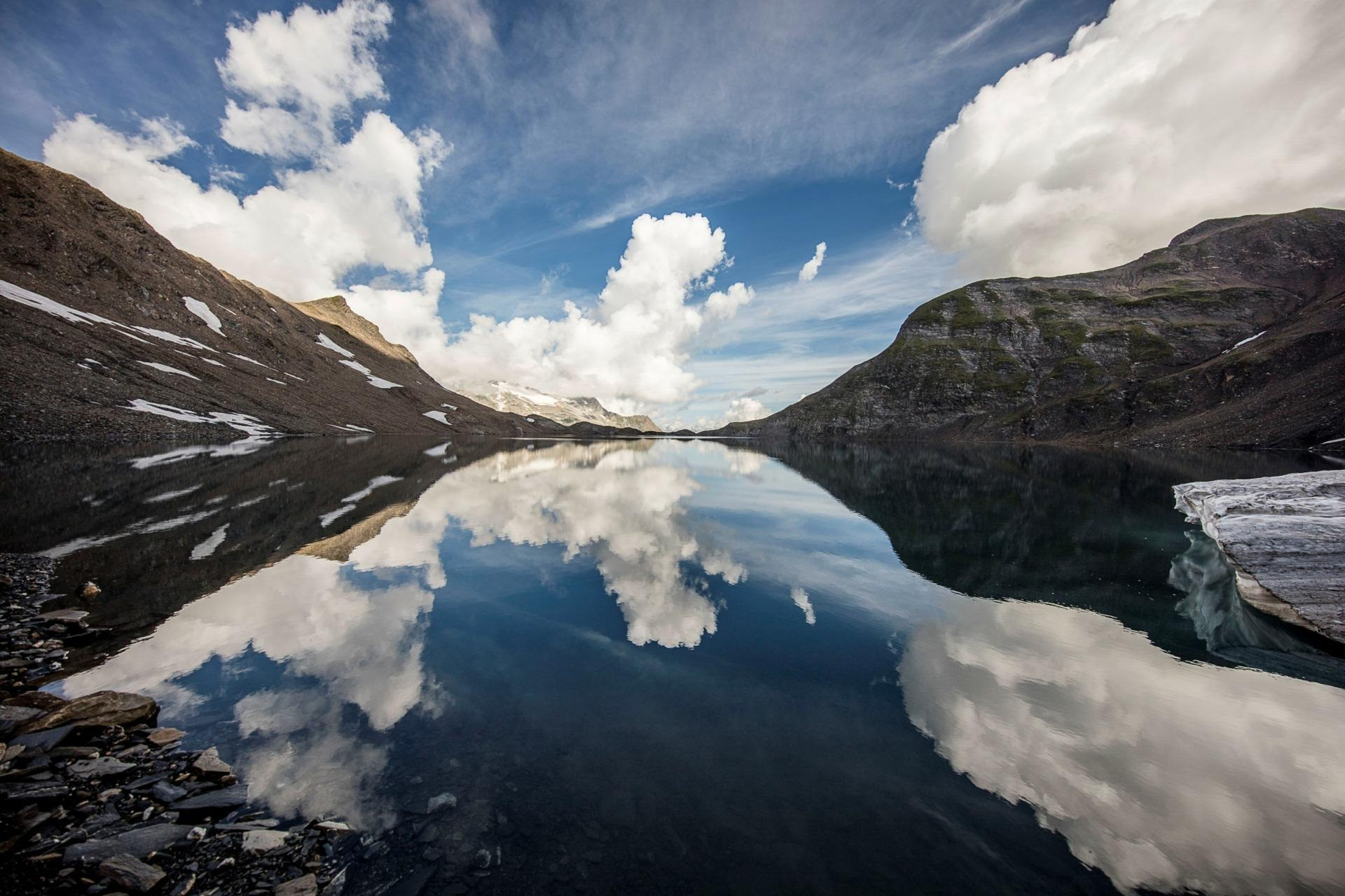 Klarer Bergsee mit Spiegelung von Wolken und umliegenden Gipfeln.