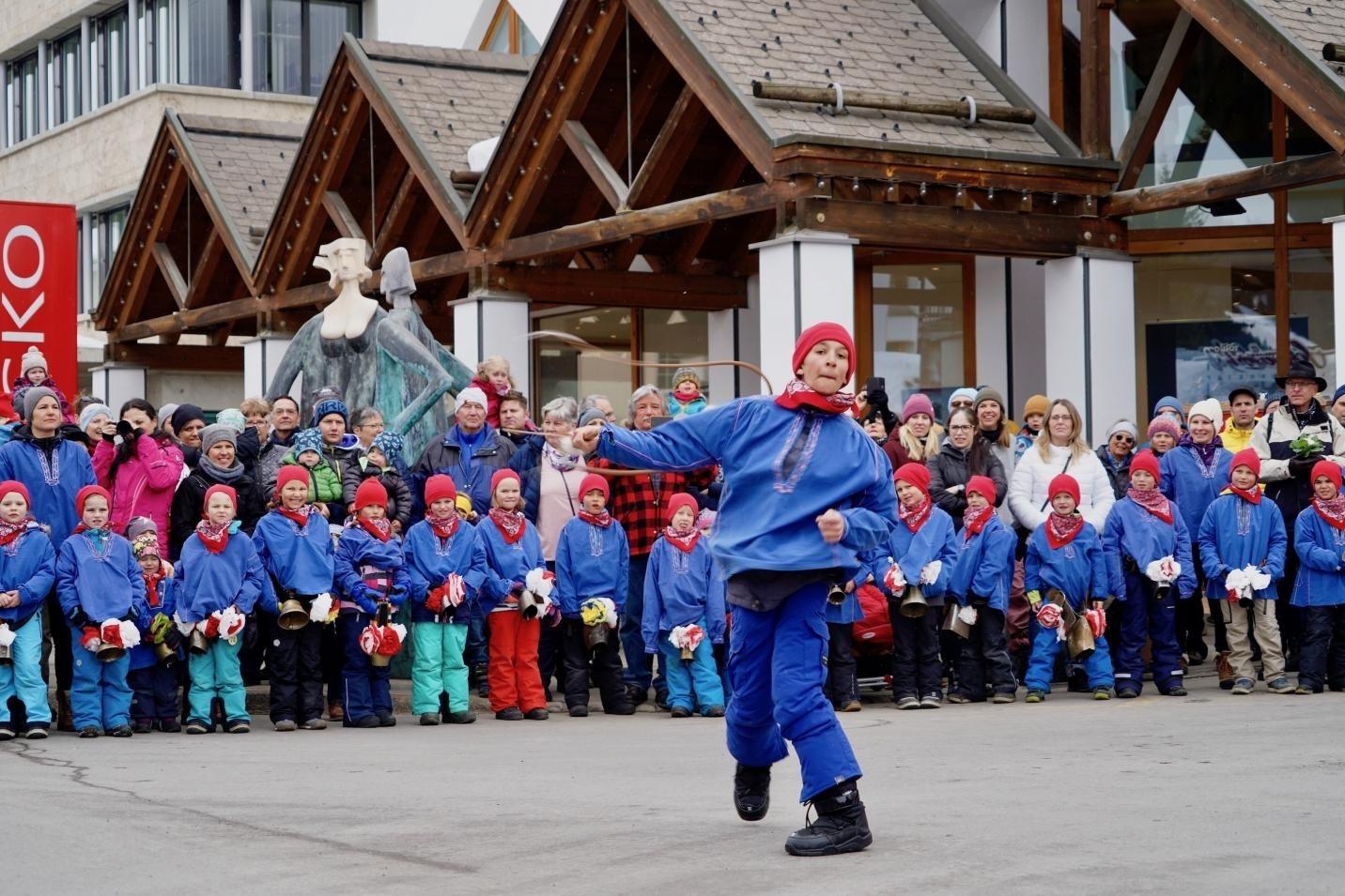 Kinderevent in Lenzerheide: Traditionelle Winteraufführung mit tanzenden Kindern in blauen Trachten und roten Mützen vor Publikum auf dem Dorfplatz.