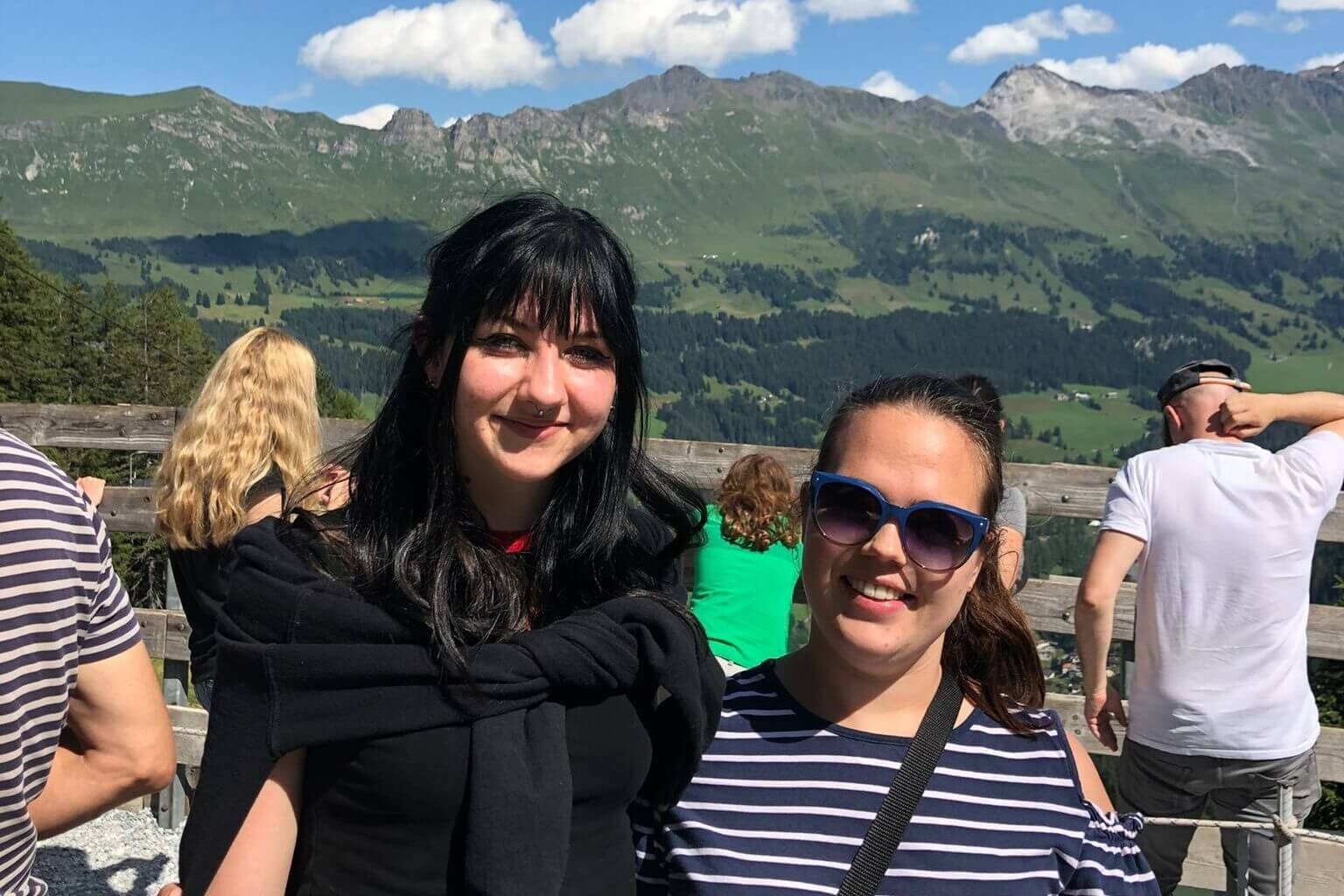 Zwei Frauen stehen draußen vor einer Berglandschaft und lächeln in die Kamera.