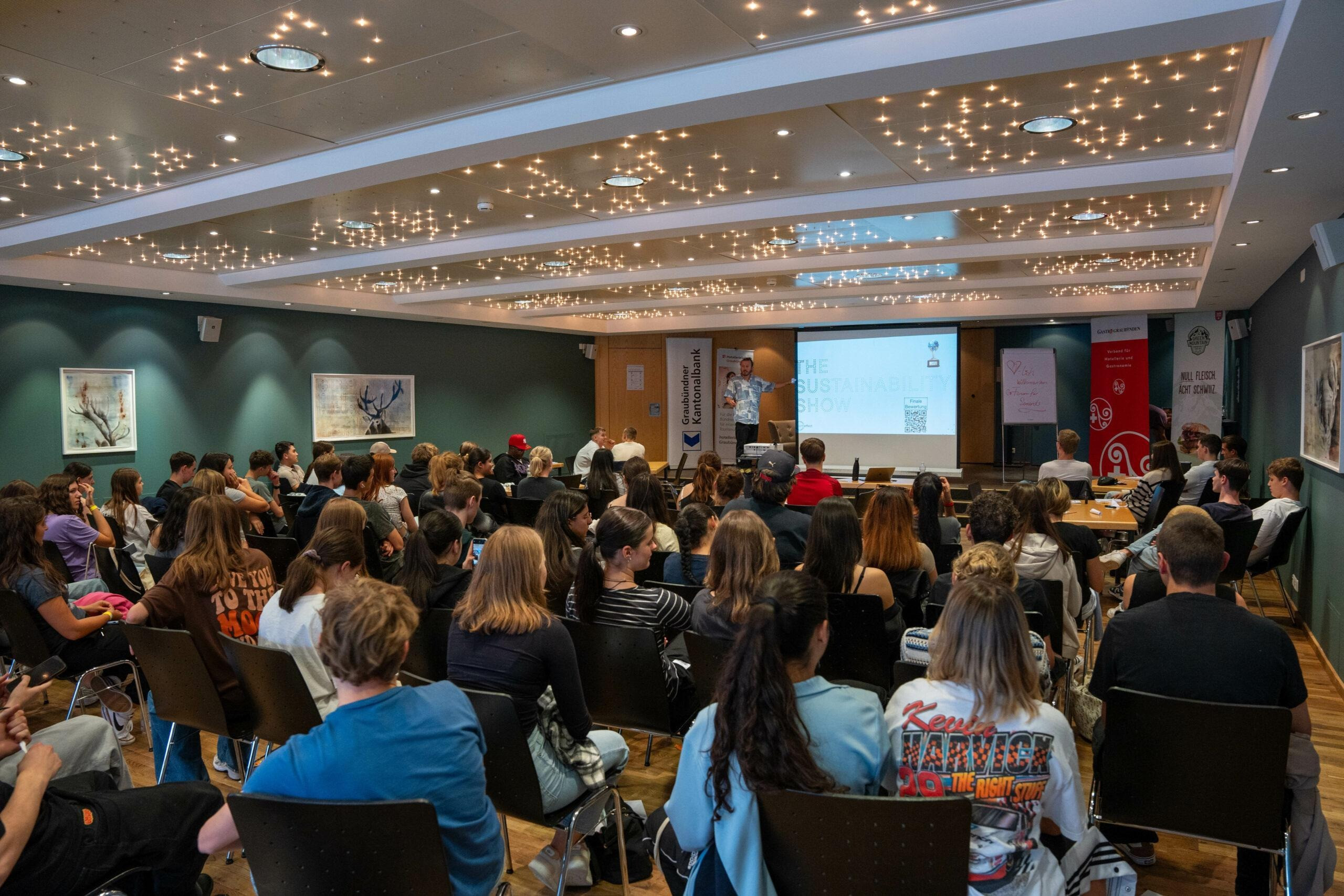 Eine grosse Gruppe junger Menschen sitzt in einem Saal und hört einer Person zu, die vorne eine Präsentation über Nachhaltigkeit hält. Der Raum ist hell beleuchtet und es hängen Bilder an den Wänden.