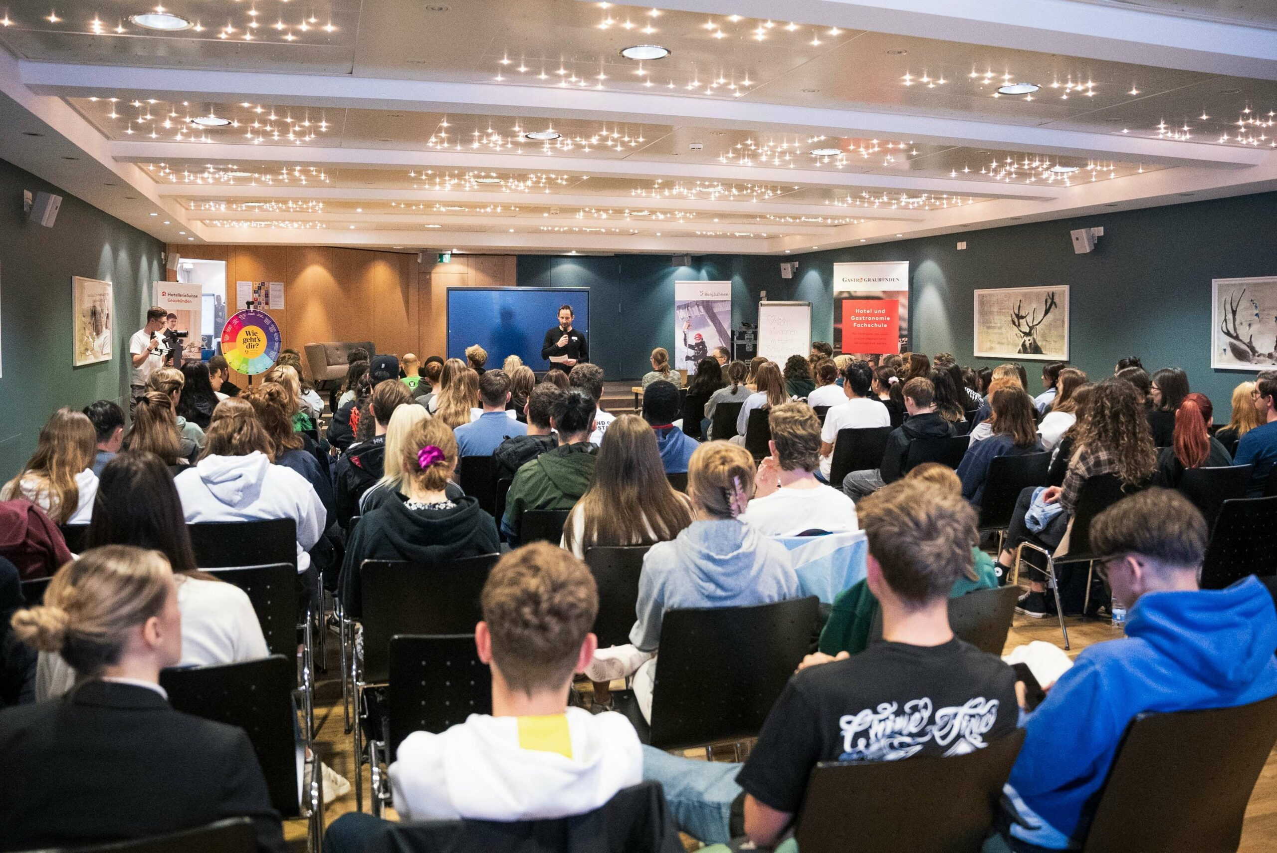 „Viele junge Menschen sitzen in einem Saal und hören einer Person auf der Bühne zu.