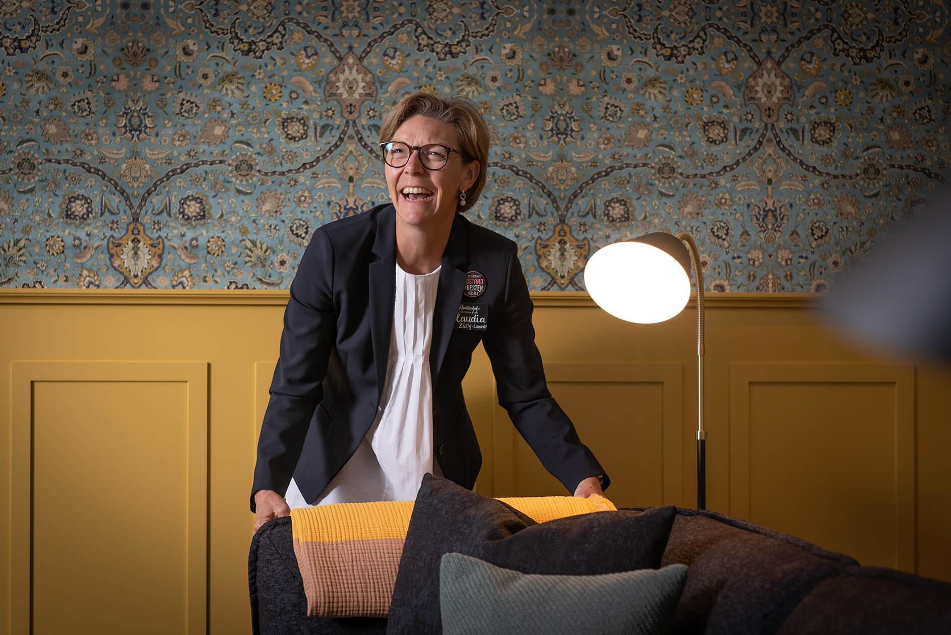 Eine lächelnde Frau in einer Hotelfach-Uniform richtet Kissen auf einem Sofa her.