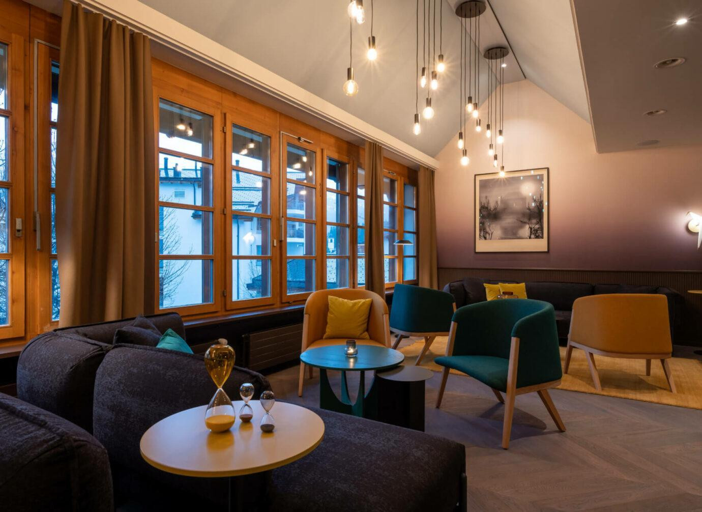 Ein gemütlicher Raum im Hotel, modern eingerichtet mit Sesseln, Sofas und vielen kleinen Tischen. Warme Beleuchtung hängt von der Decke, und grosse Fenster lassen Tageslicht hinein.