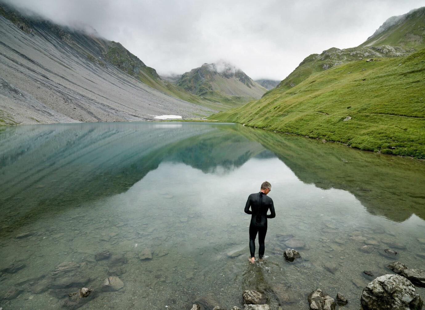 Eine Person im Neoprenanzug steht am Rand eines klaren Bergsees, umgeben von grünen Hügeln und steinigen Bergen unter bewölktem Himmel.