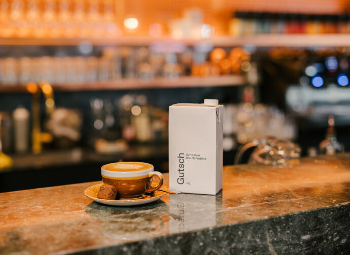 ine Tasse Kaffee mit Keks steht auf einer Marmortheke, daneben ein weißer Karton mit Haferdrink. Der Hintergrund ist unscharf mit Bar-Elementen.