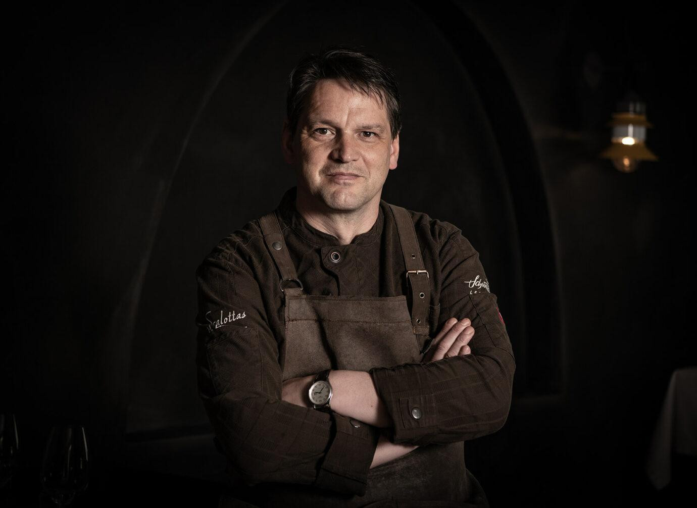 Ein Koch steht mit verschränkten Armen da. Er trägt dunkle Kochkleidung und eine Schürze. Der Hintergrund ist dunkel, und der Koch schaut direkt in die Kamera.