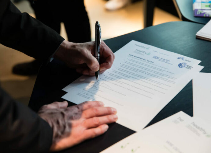 Eine Person unterschreibt mit einem Stift ein Dokument auf einem Tisch; mehrere Blätter liegen übereinander.