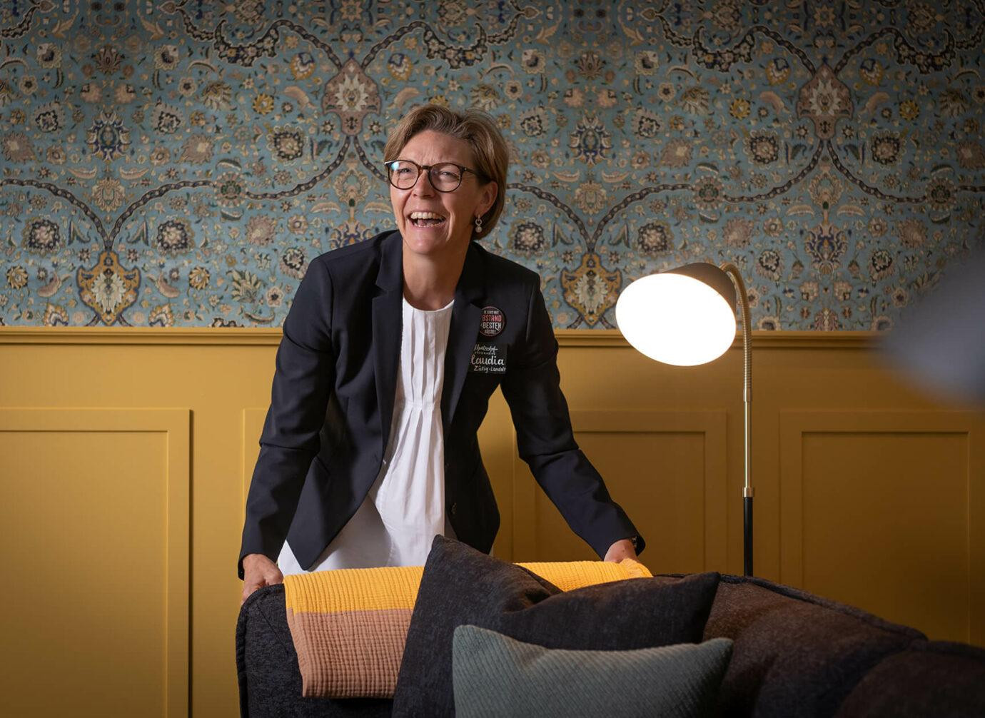 Frau mit Brille lächelt, während sie in einem gemütlichen, stilvoll dekorierten Raum eine Decke über ein Sofa legt.