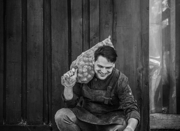 Schwarz-weiss-Foto eines lächelnden Mannes in Arbeitskleidung, der in der Hocke sitzt und einen grossen Netzsack voller Kartoffeln über der Schulter trägt. Hinter ihm steht eine Holzwand.