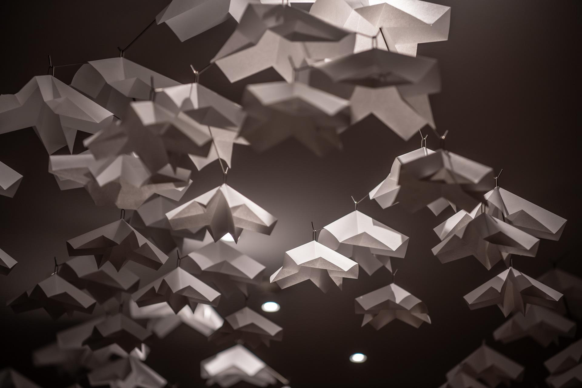 Mehrere weisse, geometrisch gefaltete Papierobjekte hängen dicht unter der Decke und erzeugen ein schwebendes, wolkenartiges Muster, das von warmem Licht sanft angestrahlt wird.