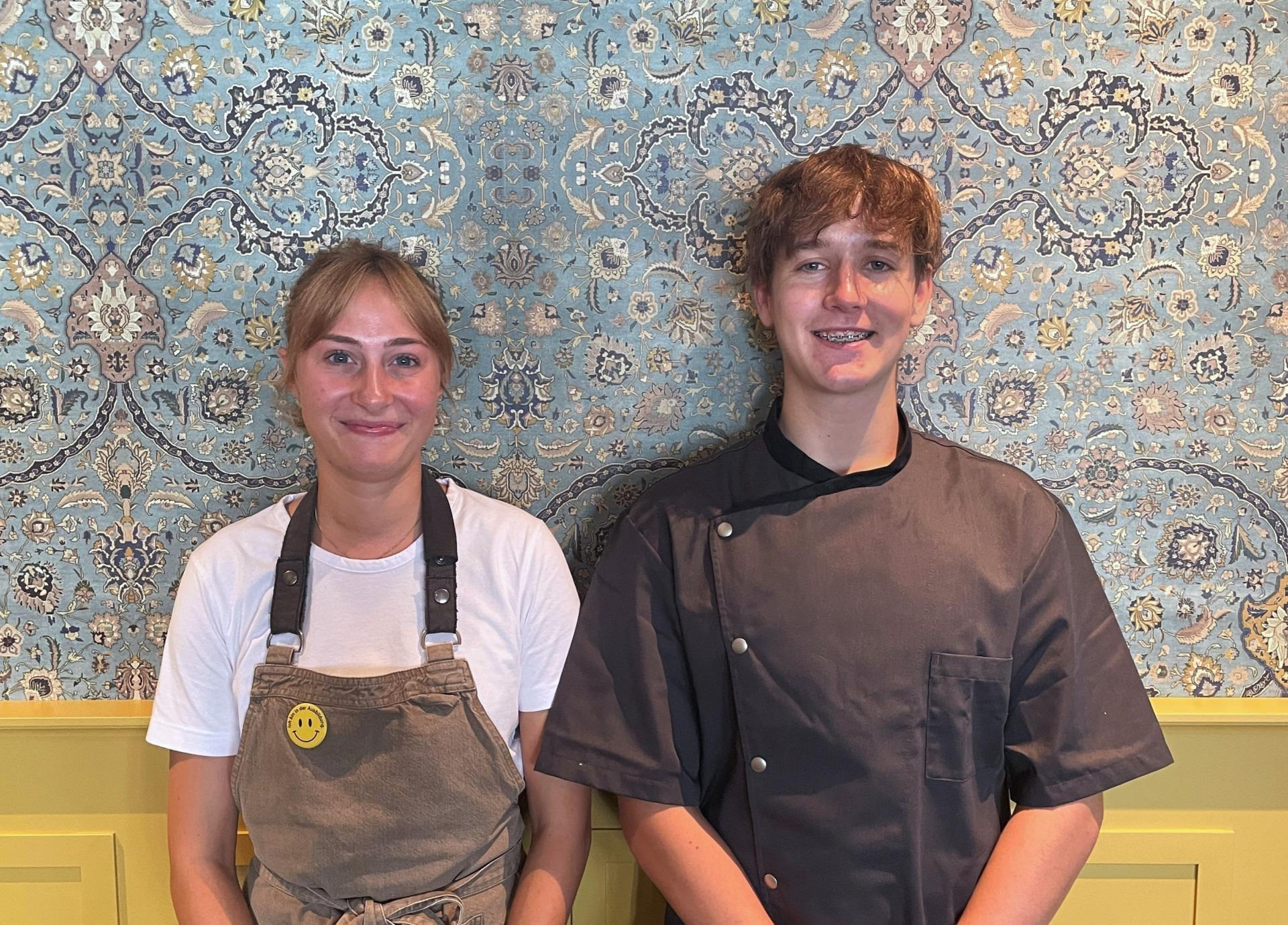 Zwei junge Mitarbeitende stehen vor einer dekorativen, blau-gold gemusterten Wand. Links eine Person mit hellem T-Shirt und Schürze mit Smiley-Anstecker, rechts eine Person in dunkler Kochjacke. Beide lächeln in die Kamera.