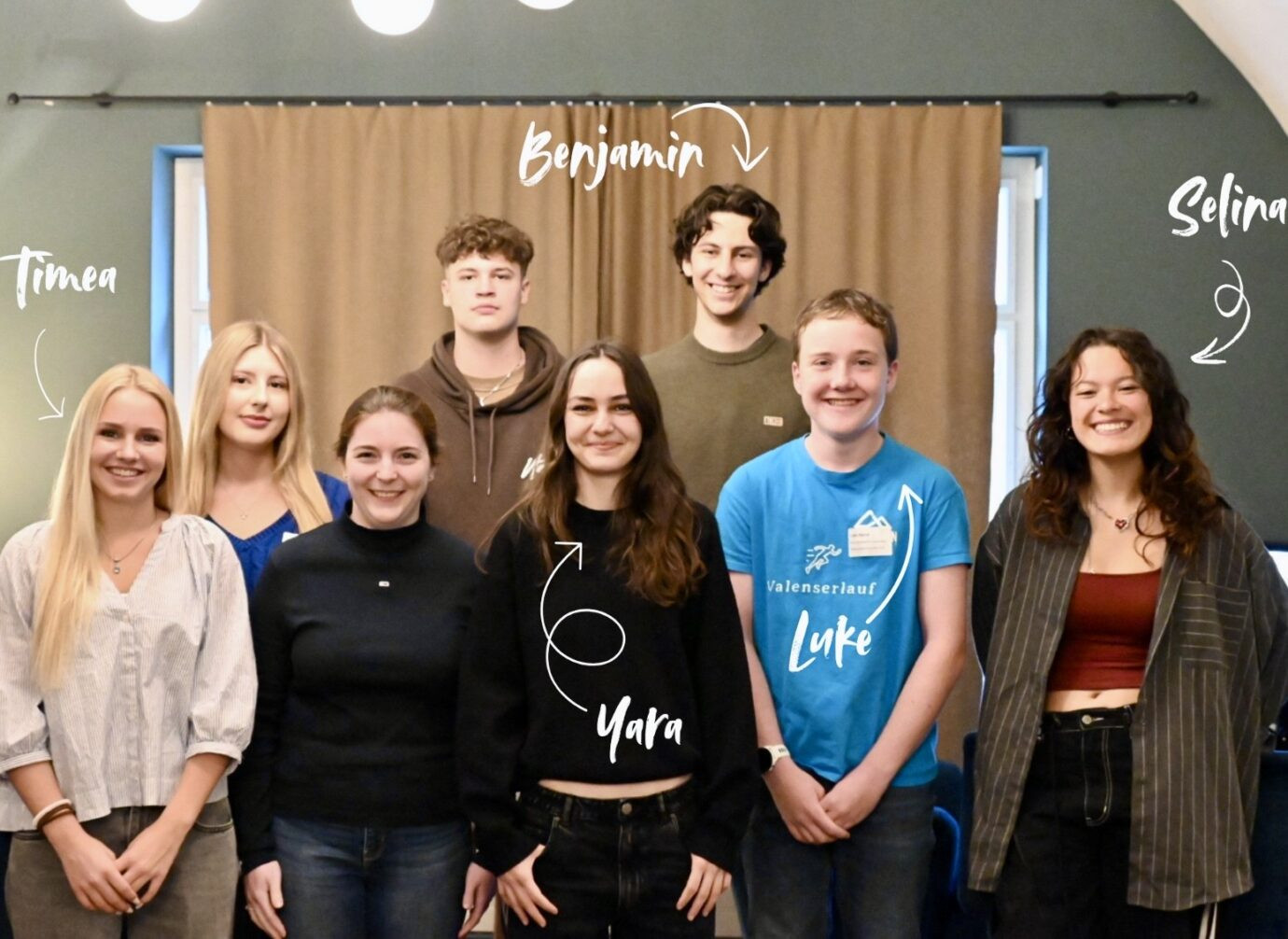Gruppe junger Menschen, die in zwei Reihen für ein Gruppenfoto posieren. Alle lächeln in die Kamera. Im Bild sind handschriftliche Namensbeschriftungen mit Pfeilen zu den jeweiligen Personen ergänzt. Die Gruppe steht vor einem beige-braunen Vorhang in einem Raum mit grünen Wänden.