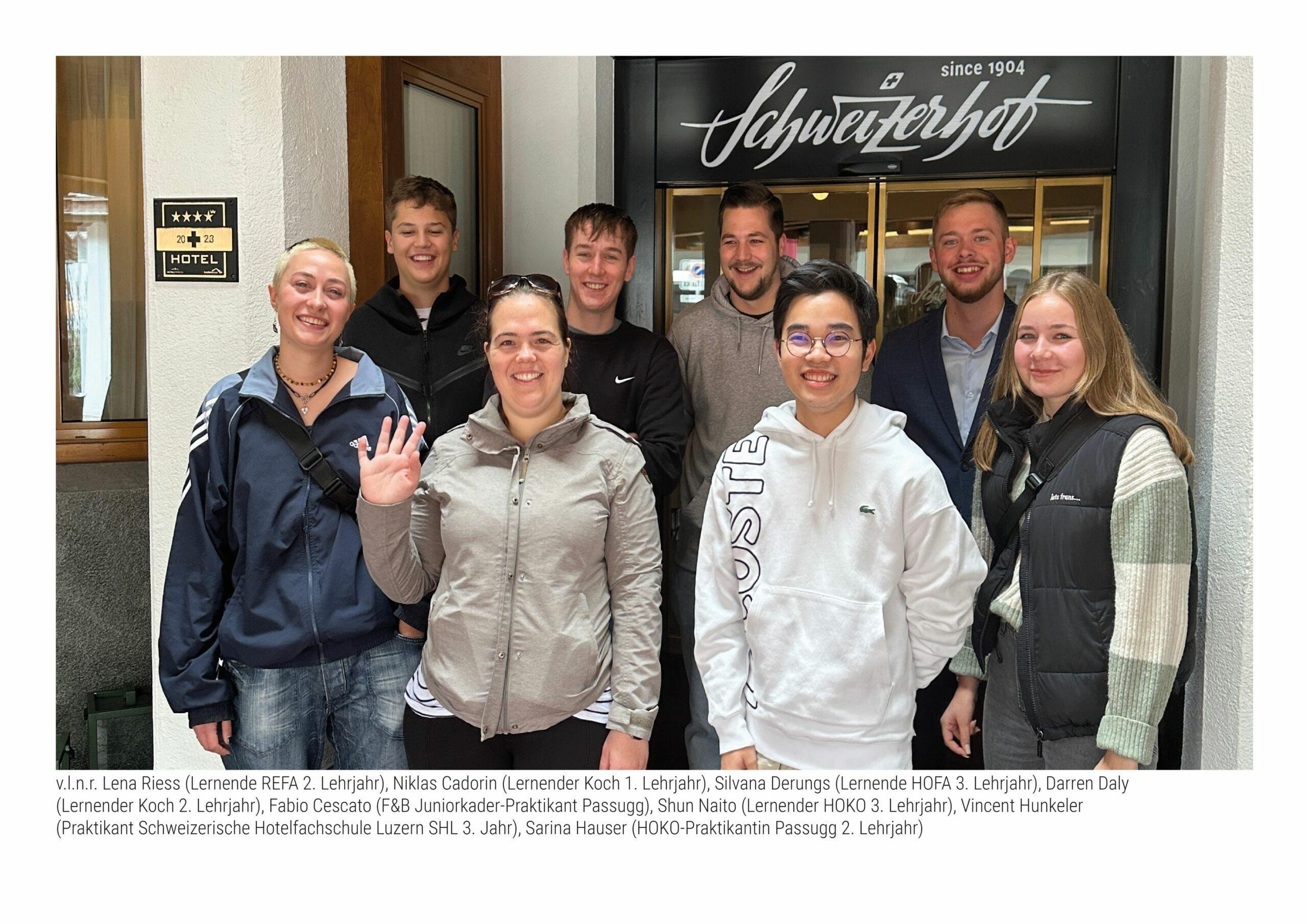Gruppe von acht jungen Erwachsenen, die lächelnd vor dem Eingang eines Hotels stehen. Hinter ihnen ist ein Schild mit vier Sternen und der Schriftzug „Schweizerhof“ über der Tür zu sehen. Die Personen tragen Freizeitkleidung oder sportliche Jacken, eine Person vorne links winkt in die Kamera.