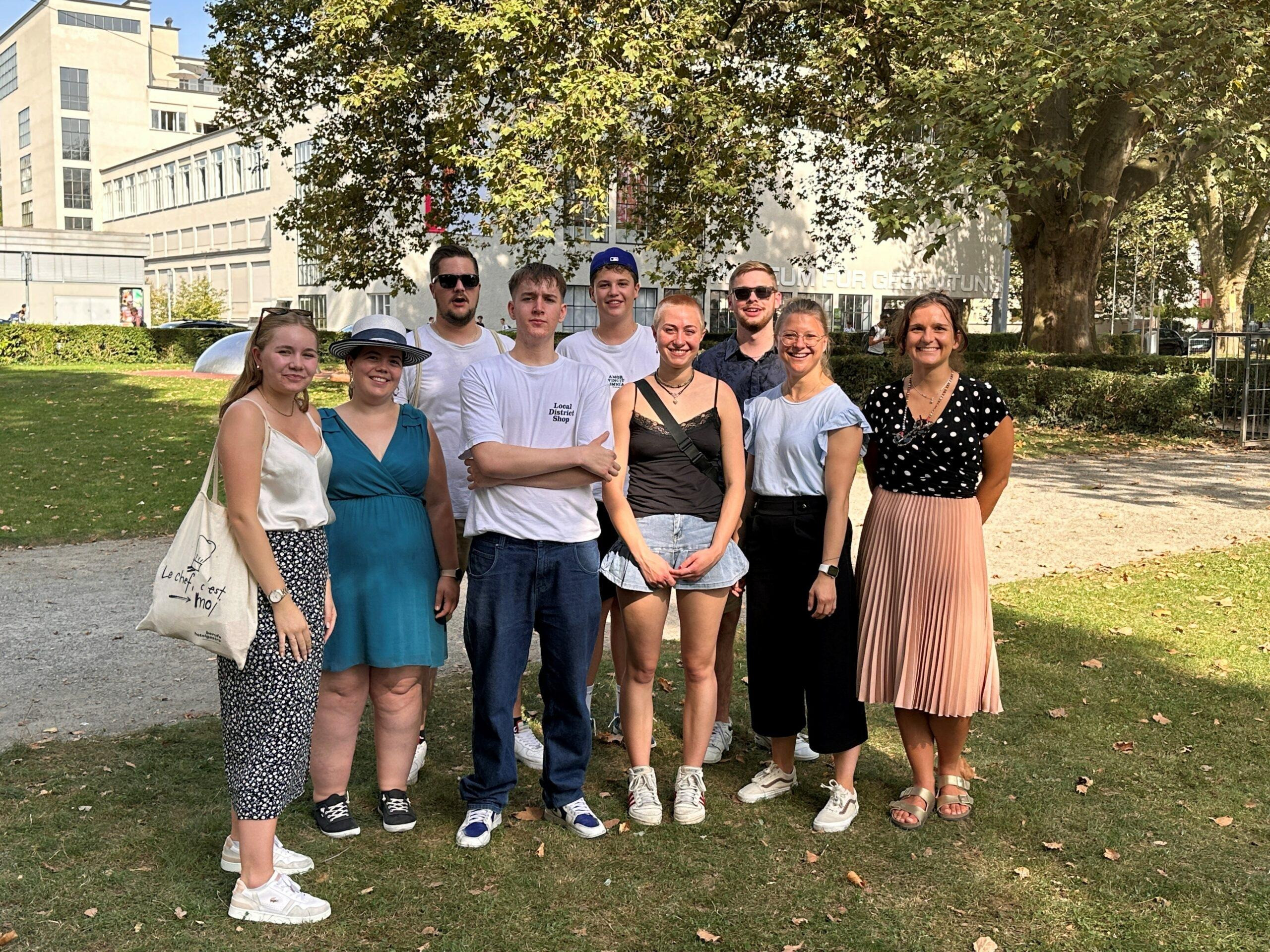 Gruppe von zehn jungen Erwachsenen, die im Freien auf einer Wiese stehen und in die Kamera lächeln. Einige tragen sommerliche Kleidung wie Shorts, Röcke oder leichte Tops. Im Hintergrund sind Bäume, ein Gebäude und ein sonniger Himmel zu sehen, was eine entspannte Ausflugsstimmung vermittelt.