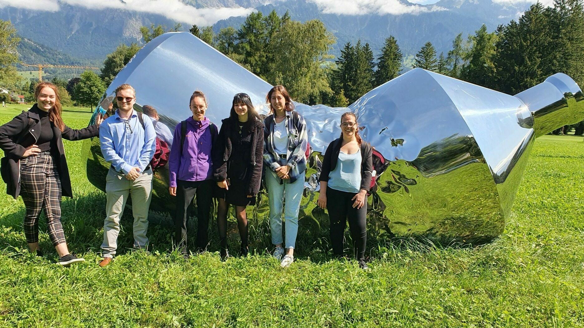 Sechs Personen stehen auf einer Wiese vor einer grossen, spiegelnden Metallskulptur, die wie ein überdimensioniertes, glänzendes Gefäss oder Trichter geformt ist. Im Hintergrund sind Bäume, Wiese und hohe Berge unter leicht bewölktem Himmel zu sehen.