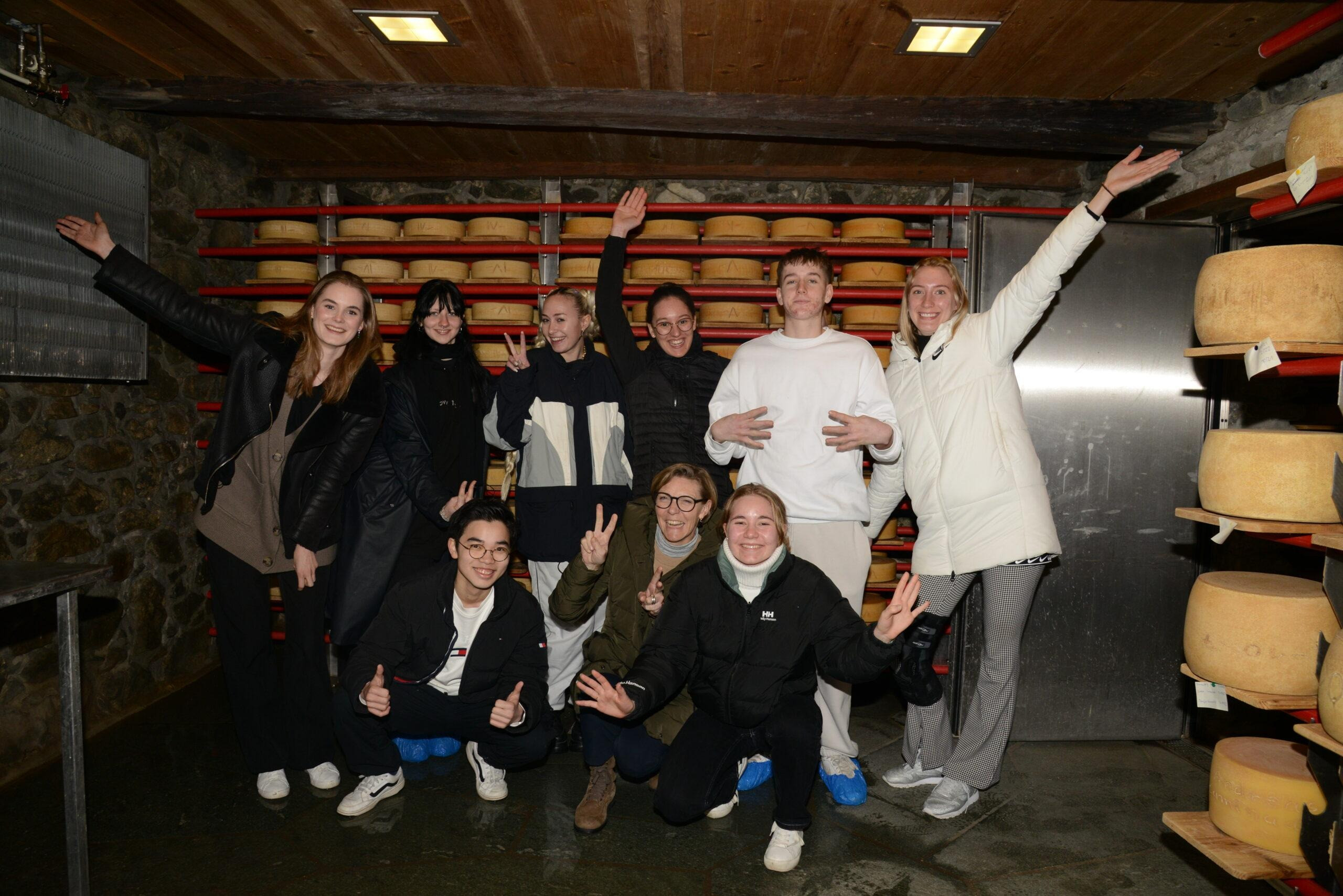Eine fröhliche Gruppe von zehn Personen posiert in einem Käselager, umgeben von grossen runden Käselaiben auf Regalen. Alle tragen warme Kleidung, einige machen Peace-Zeichen oder strecken die Arme in die Höhe. Die Stimmung wirkt ausgelassen und fröhlich.