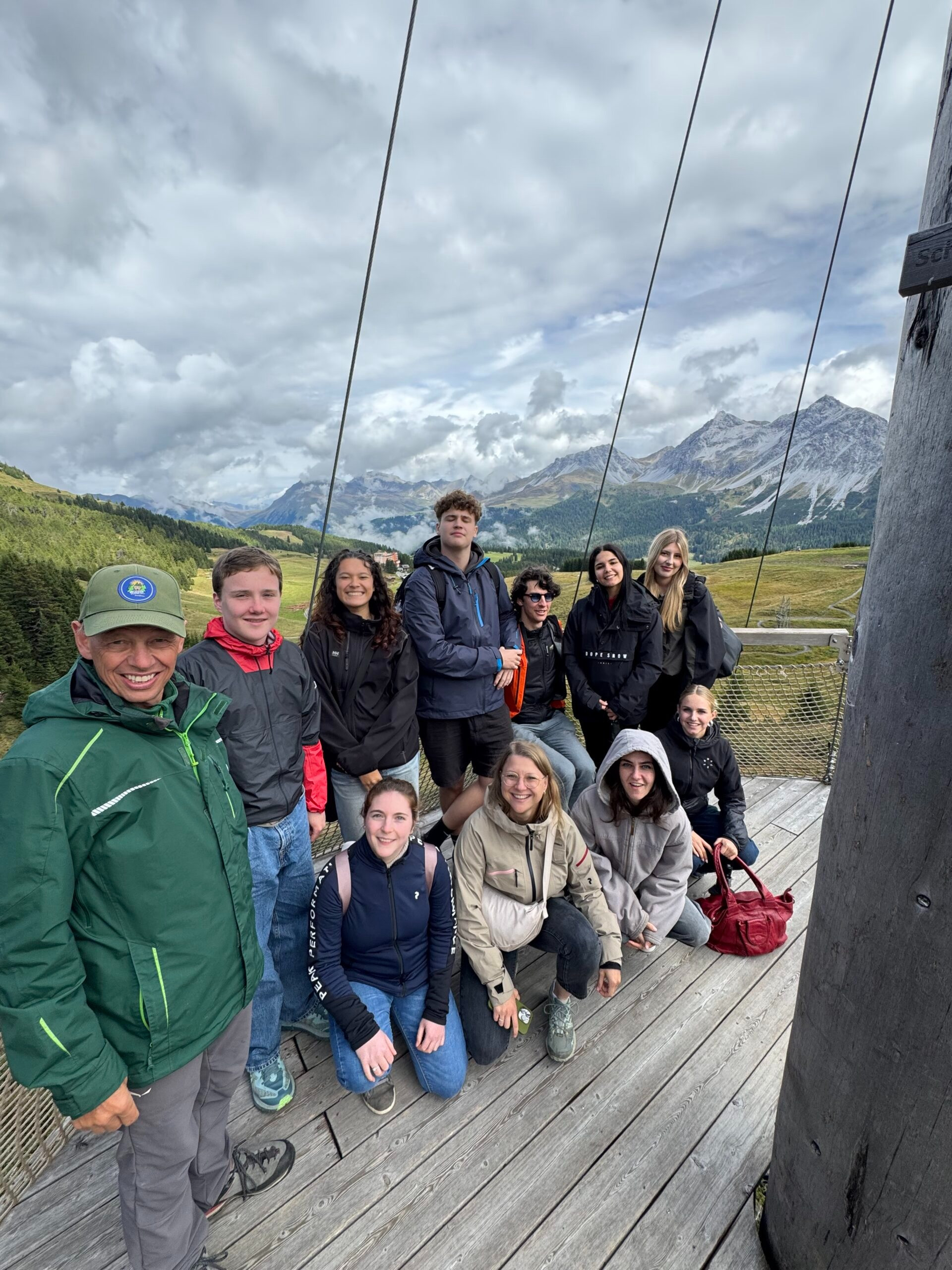 Gruppe von elf Personen in Outdoor-Kleidung steht und sitzt auf einer Holzplattform im Gebirge. Hinter ihnen erstreckt sich eine weite Berglandschaft mit grünen Hängen, Wald und hohen Gipfeln unter einer bewölkten, dramatischen Himmelkulisse. Alle lächeln und wirken, als wären sie auf einer gemeinsamen Wanderung oder Exkursion.