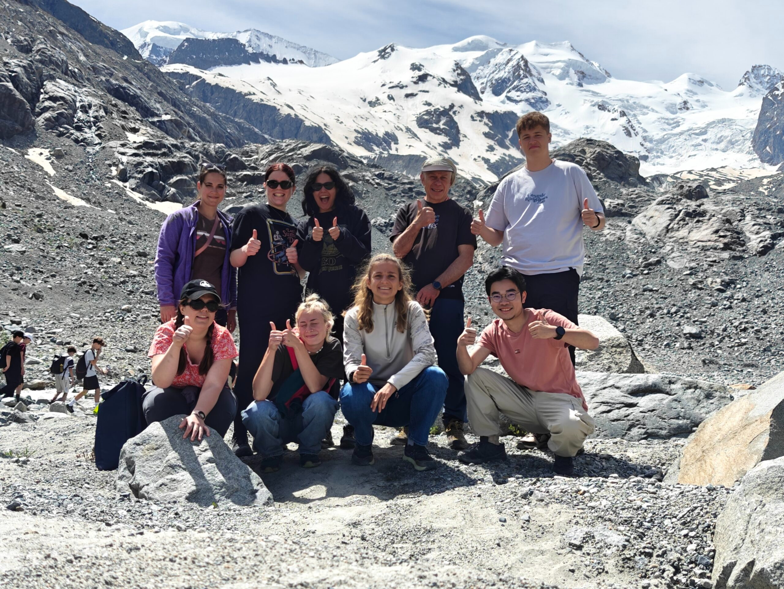 Gruppe von zehn Personen posiert fröhlich vor einer eindrucksvollen Gletscher- und Bergkulisse. Alle zeigen Daumen hoch oder lachen in die Kamera. Sie stehen und hocken auf felsigem Untergrund, umgeben von grauen Steinen, Schneefeldern und hohen, schneebedeckten Gipfeln unter blauem Himmel.