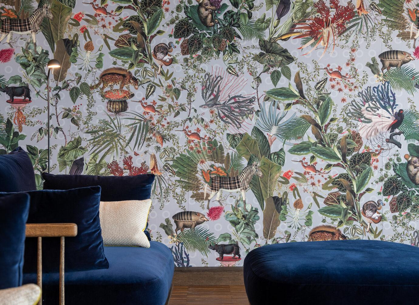 ChatGPT: Blick in einen gemütlichen Lounge-Bereich mit dunkelblauen Polstermöbeln vor einer auffälligen, farbenfrohen Tapete im Dschungelstil. Die Wand zeigt ein dichtes Muster aus Pflanzen, Blättern und exotischen Tieren wie Zebras, Leoparden und Vögeln. Eine schlichte Stehleuchte ergänzt die lebendige, verspielte Atmosphäre.