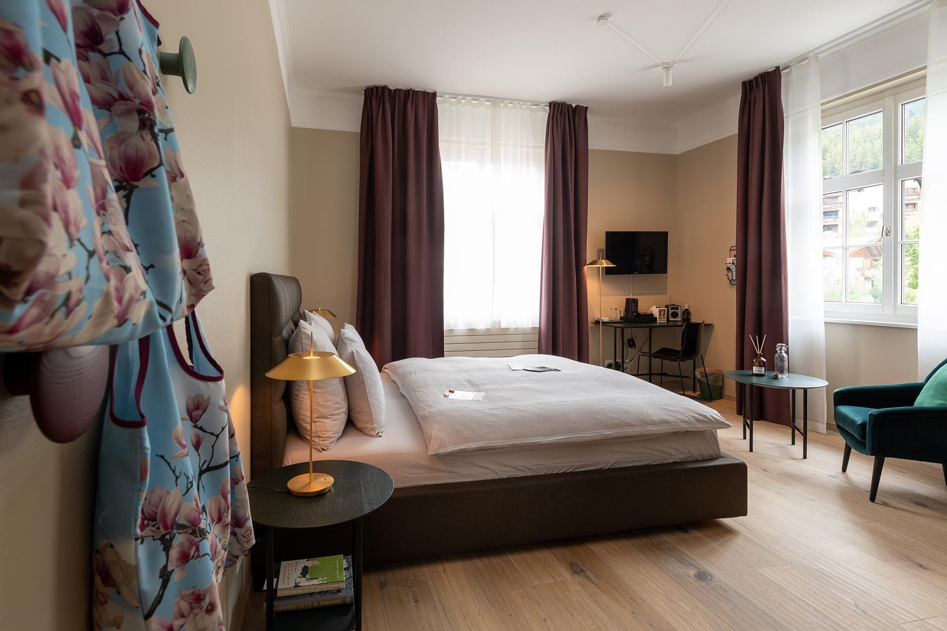 Hotelzimmer mit grossem Bett und Fenster mit langen Vorhängen. Ein Beistelltisch mit Duftstäbchen und Wasserfalsche neben