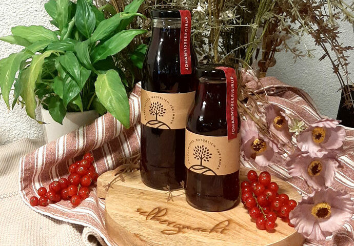 Sirup in Gläsern die auf einem Holzbrett stehen umgeben von Blüten und Johannisbeeren.