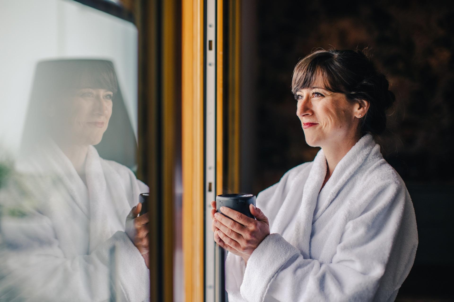 Frau steht vor einem Fenster mit einer Tasse in der Hand im Bademantel im Spabereich.
