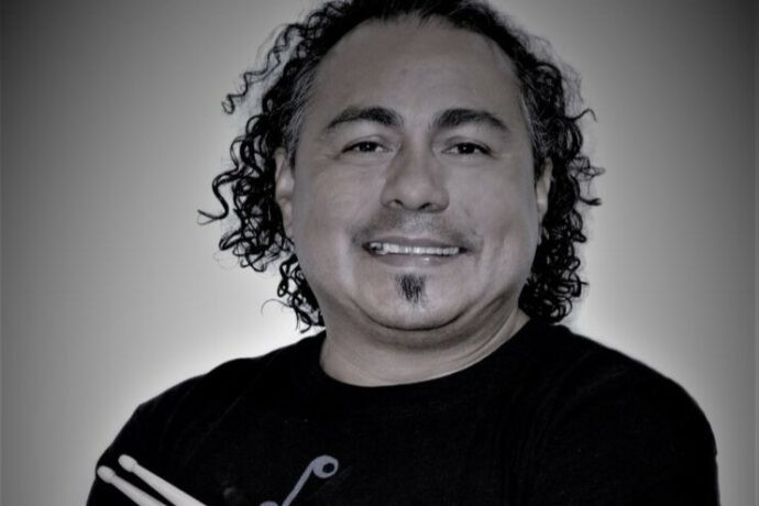 **Alternativtext:** Ein Mann mit schulterlangen, lockigen Haaren lächelt in die Kamera. Er trägt ein Oberteil mit einem aufgedruckten Symbol und hält ein Paar Schlagzeugstöcke im Arm. Seine Gesichtsbehaarung umfasst einen kleinen Bartfleck unter der Unterlippe.