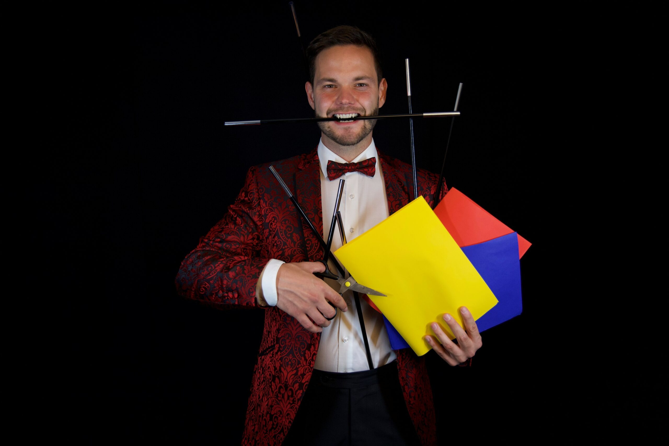 Ein Mann in einem roten, gemusterten Anzug und weissem Hemd steht vor schwarzem Hintergrund. Er hält mehrere bunte Papierblätter in Gelb, Blau und Rot in der Hand. In derselben Hand stecken mehrere schwarze Stäbe, die kreuz und quer abstehen. Zusätzlich hält er eine grosse Schere, und ein weiterer Stab ragt quer aus seinem Mund.