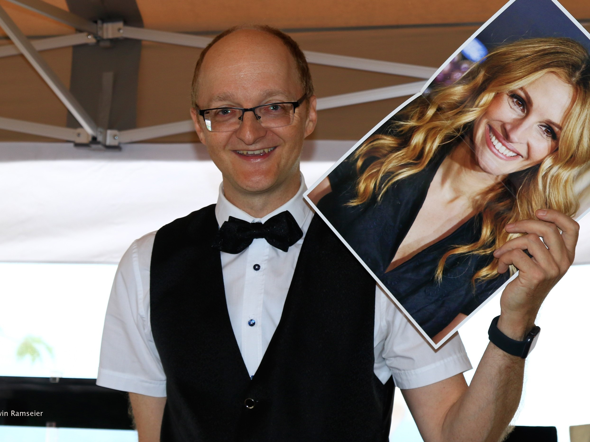 Ein Mann mit Brille, weissem Hemd, schwarzer Weste und schwarzer Fliege steht unter einem Zeltdach. In der rechten Hand hält er ein grosses Foto einer lächelnden Frau mit langen blonden Haaren.