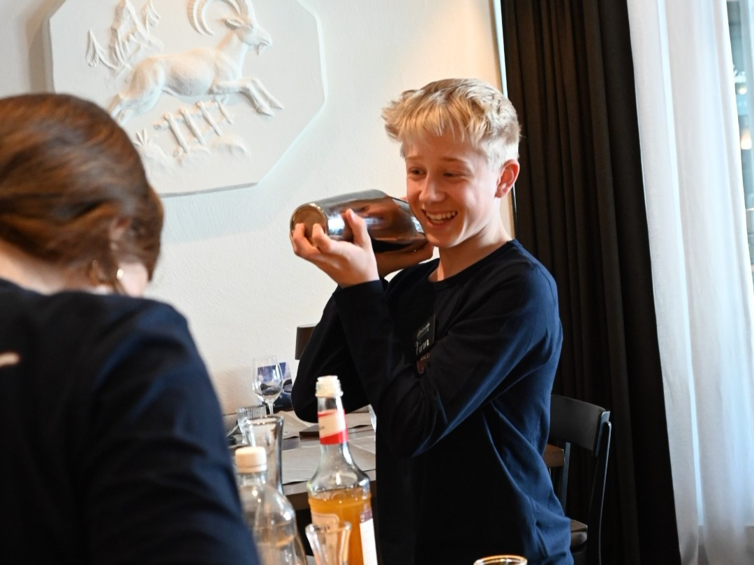 Ein Junge mit blondem Haar und dunkelblauem Langarmshirt steht lachend an einem Tisch und schüttelt einen Metallshaker. Vor ihm stehen verschiedene Flaschen und Gläser. Links im Vordergrund ist eine weitere Person teilweise zu sehen.