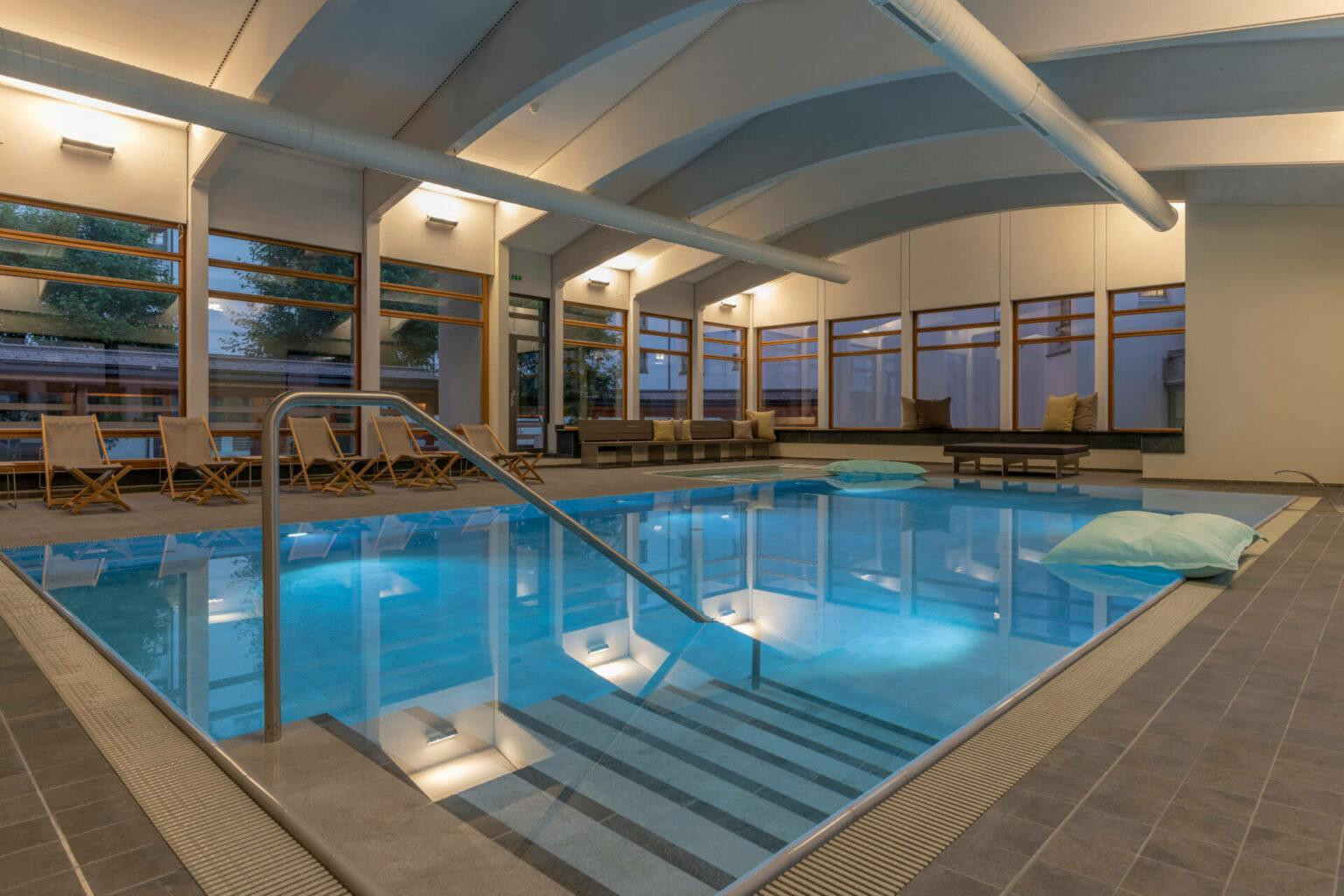 Ein Innenraum mit einem rechteckigen Becken. Ueber eine breite Treppe mit Geländer gelangt man ins Wasser. Rund um das Becken stehen mehrere Klappstuehle und Sitzbaenke mit Kissen. Grosse Fenster reichen bis fast zum Boden und zeigen Baeume draussen. Auf der Wasseroberflaeche treiben zwei grosse Kissen. Die Decke ist gebogen und von langen Rohren und Lampen durchzogen.