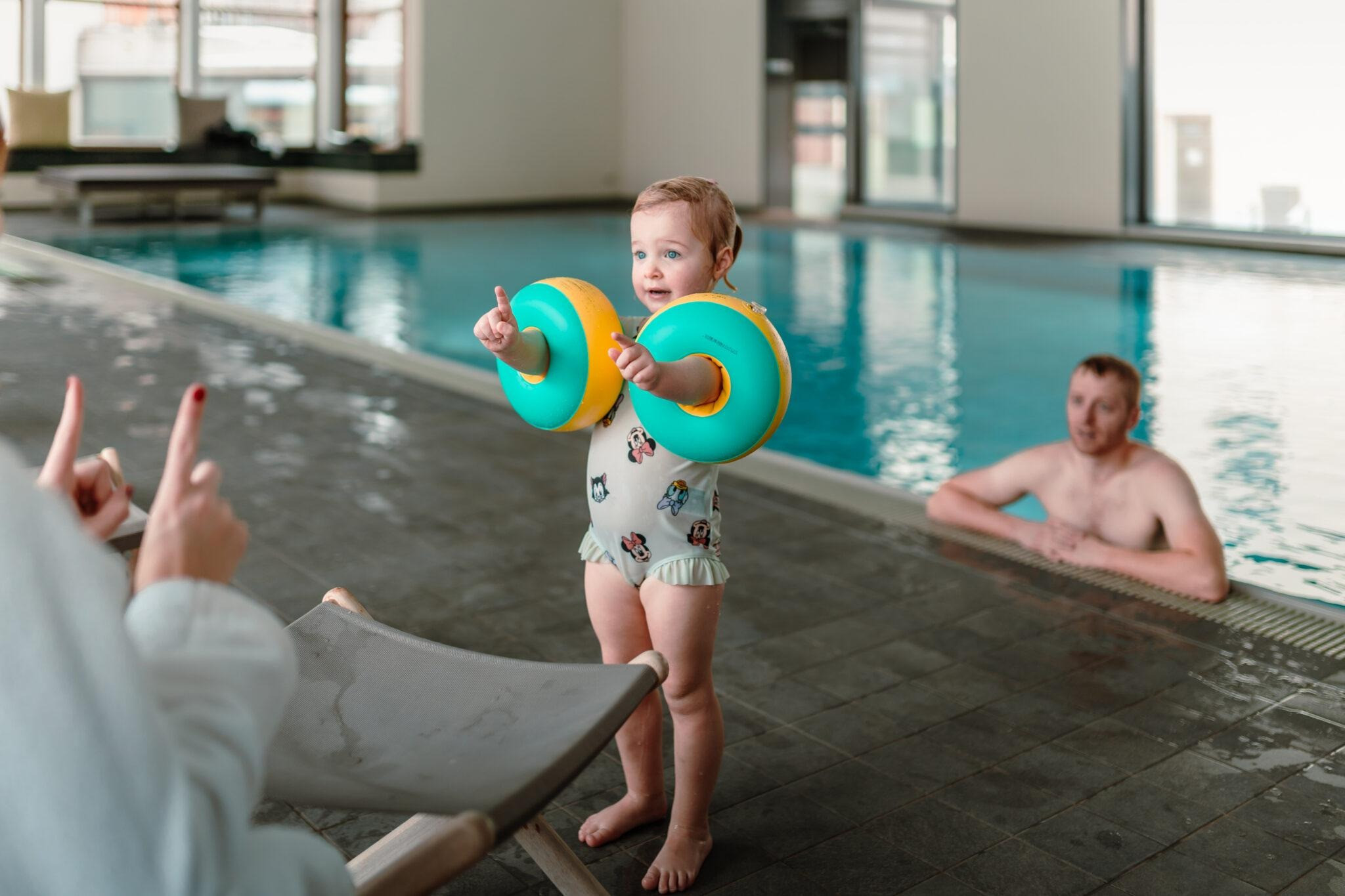 Ein Kleinkind mit grossen Schwimmringen an den Armen steht am Beckenrand und zeigt mit beiden Haenden nach vorn. Eine erwachsene Person sitzt im Wasser und schaut zum Kind hin.