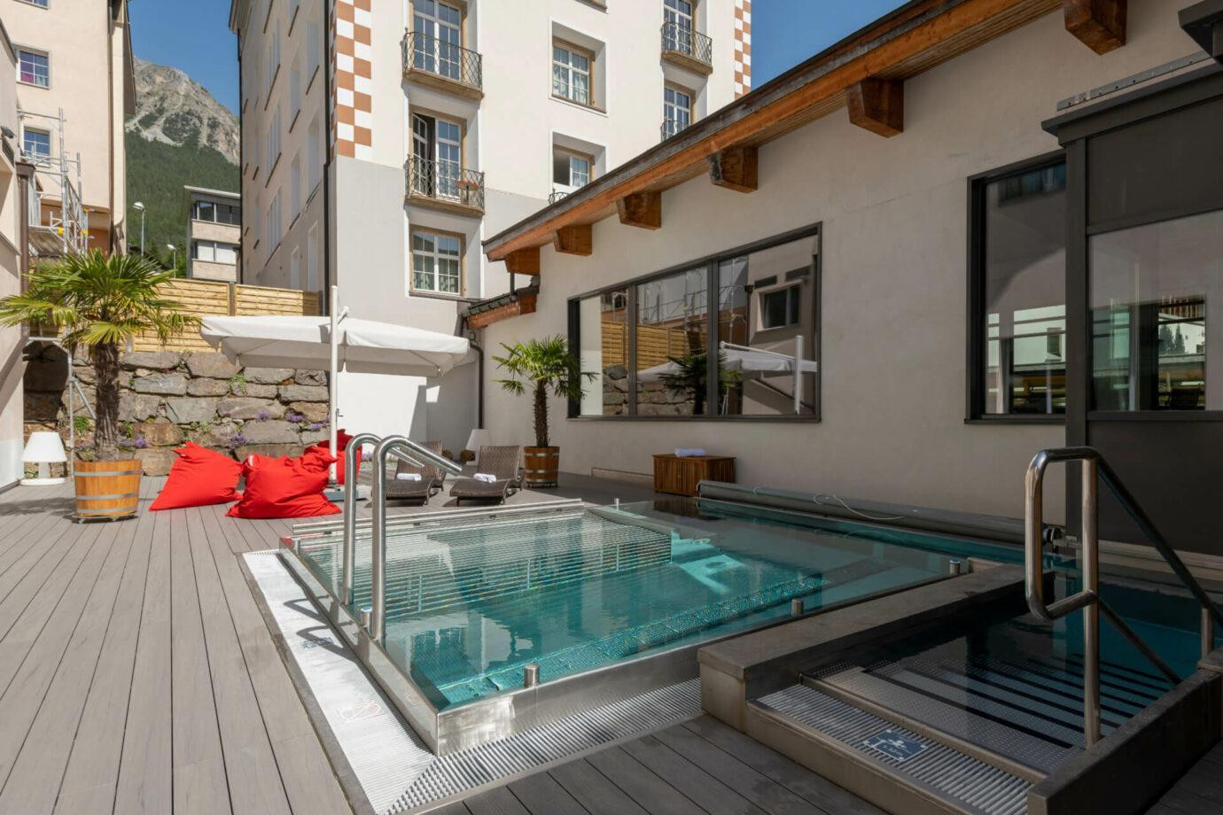 Ein kleiner Aussenbereich mit einem rechteckigen Whirlpool und einer schmalen Treppe ins Wasser. Rundherum stehen Liegestuehle, grosse Sitzsaecke und zwei Topfpalmen. Der Bereich ist von hohen Gebaeuden umgeben; im Hintergrund ragt ein steiler Berg auf.