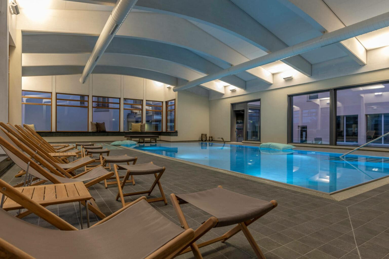 Ein Innenraum mit einem rechteckigen Becken. Ueber eine breite Treppe mit Geländer gelangt man ins Wasser. Rund um das Becken stehen mehrere Klappstuehle und Sitzbaenke mit Kissen. Grosse Fenster reichen bis fast zum Boden und zeigen Baeume draussen. Auf der Wasseroberflaeche treiben zwei grosse Kissen. Die Decke ist gebogen und von langen Rohren und Lampen durchzogen.