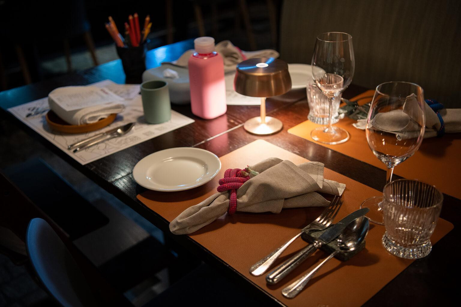 Ein gedeckter Restauranttisch mit Geschirr, Besteck, Gläsern und Stoffservietten. Auf dem Tisch liegen auch Kinderutensilien wie eine Trinkflasche, ein Becher und Stifte. Eine kleine Tischlampe beleuchtet die Szene.