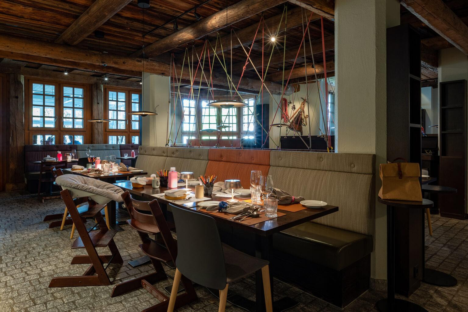 Ein Restaurantbereich mit langen Sitzbänken, Holztischen und mehreren eingedeckten Plätzen. Auf den Tischen stehen Gläser, Teller, Besteck sowie Kinderutensilien wie Trinkflaschen und Stifte. Im Raum sind grosse Holzdeckenbalken und Fenster mit Tageslicht sichtbar.