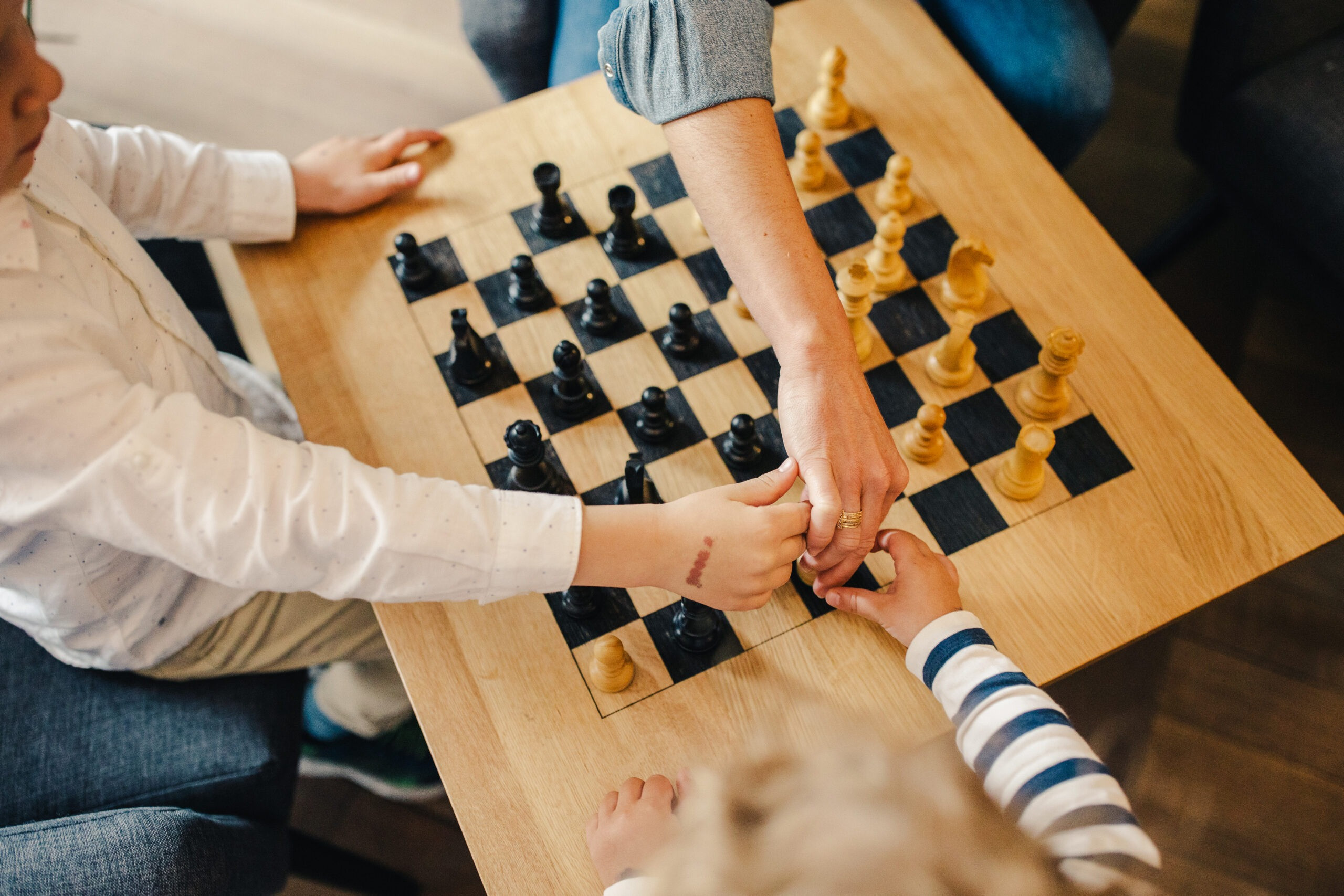 Kinder und eine erwachsene Person spielen gemeinsam Schach an einem Holztisch.