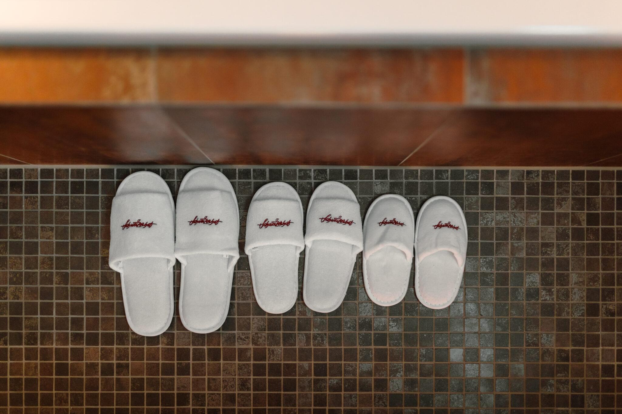 Drei Paar weisse Hausschuhe in verschiedenen Grössen ordentlich auf einem dunklen Mosaikboden ausgerichtet.