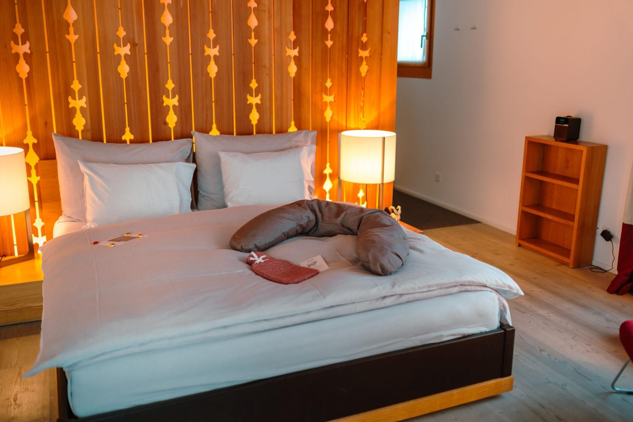 Gemütliches Hotelzimmer mit grossem Doppelbett, warm beleuchteter Holzrückwand und einfachen Holzmöbeln. Auf dem Bett liegen ein Stillkissen und kleine Willkommensgeschenke.