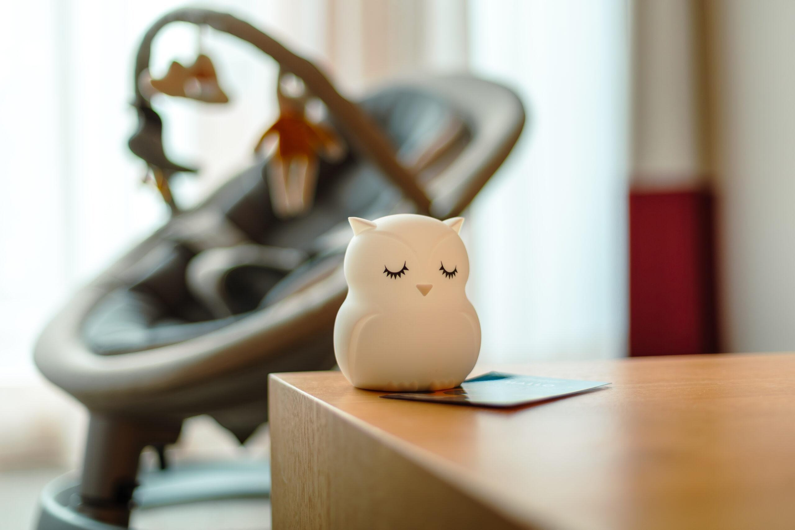 Kleine Eulenfigur mit geschlossenen Augen auf einem Holztisch, dahinter ein unscharfer Babywipper.