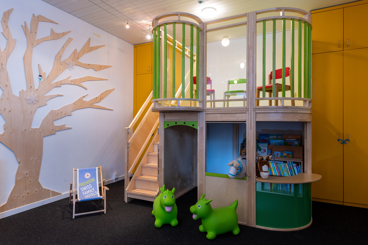 Spielzimmer mit Holzpodest, kleiner Treppe, bunten Kinderstühlen, zwei grünen Hüpfspielzeugen und einer Bücher- und Spielecke.