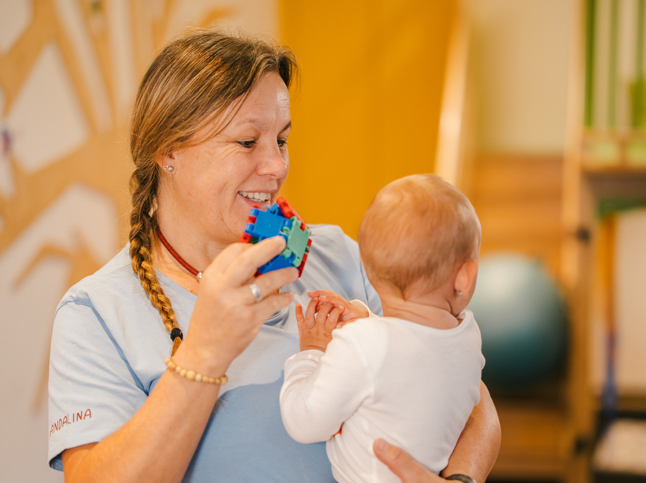 Betreuerin hält ein Baby im Arm und zeigt ihm ein buntes Spielzeug.