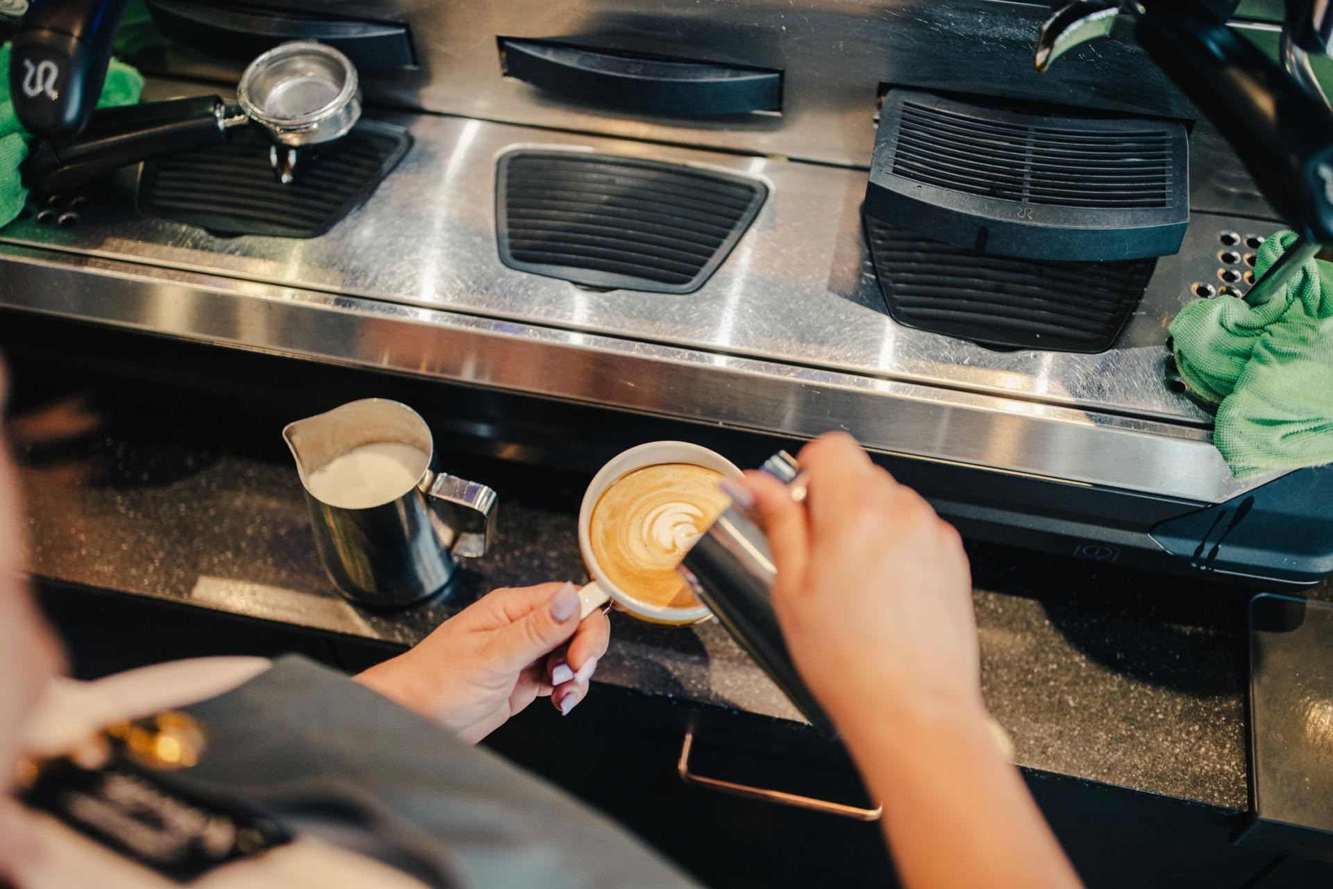 Nahaufnahme von einer Mitarbeiterin die Milch in den Kaffee giesst und vor der Kaffeemaschiene steht.