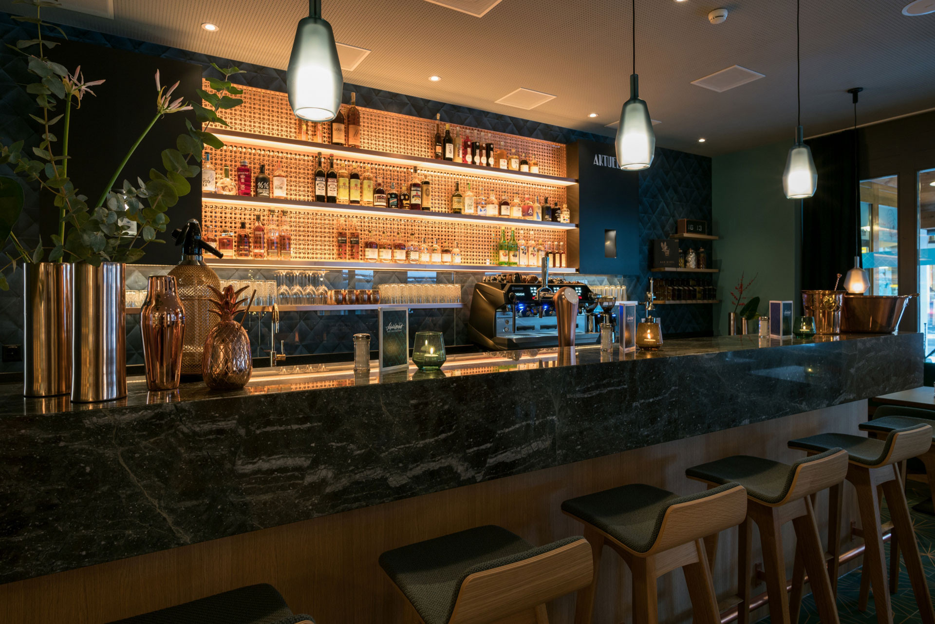 Moderne Hotelbar mit dunkler Marmor-Theke, hochwertigen Holzhockern, warm beleuchteten Regalen voller Spirituosen und gemütlicher Abendstimmung im Hotel Schweizerhof in Lenzerheide.