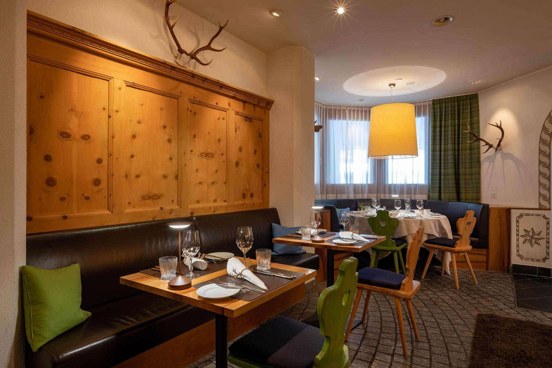 Holzgetäfeltes, gemütliches Restaurant mit dunkler Sitzbank, gedeckten Tischen, grünen Stühlen und Hirschgeweihen an der Wand.
