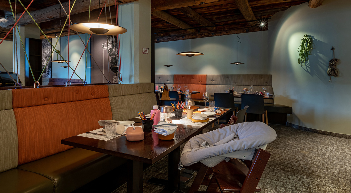 Restaurantbereich mit gedeckten Tischen, Sitzbänken in verschiedenen Farben, Babyhochstuhl im Vordergrund und dekorativen Kletterseilen an Decke und Wänden.